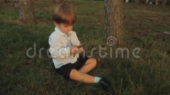 一个穿着黑色马裤的小男孩一件带领带的白色衬衫坐在绿草上玩着智能手机视频的预览图