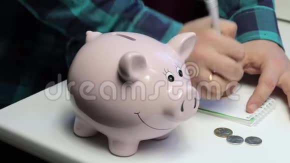 桌子上有一只粉红色的猪形的储蓄罐一个人拿着硬币把它们扔进储蓄罐做笔记视频的预览图