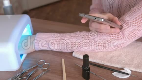 女人在紫外线灯下用紫胶擦干指甲在手机上浏览互联网特写的手视频的预览图