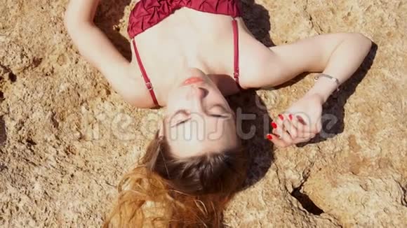 年轻的金发女孩穿着红色泳衣在岩石上晒太阳视频的预览图
