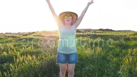 戴帽子的快乐年轻女子在夕阳下的绿野中享受夏日视频的预览图