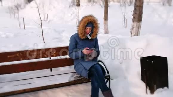 白天在下雪的天气里女人坐在长椅上在冬天的城市公园里浏览手机视频的预览图