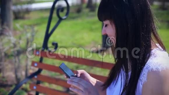 东方美丽的女孩穿着白色衬衫坐在公园的长凳上拿着智能手机笑着情绪视频的预览图