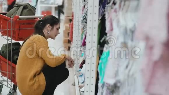 年轻漂亮的女人蹲在超市在橱窗里挑选婴儿服装靠近女性购物车4K视频的预览图