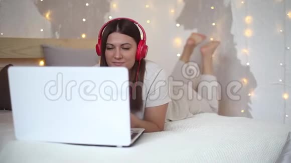 穿着睡衣的漂亮女孩用笔记本电脑在耳机上听音乐心情很好躺在床上视频的预览图