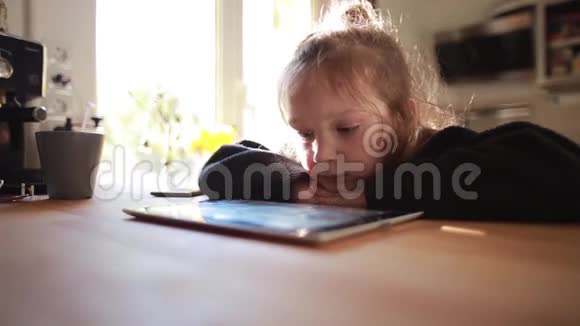 可爱的小女孩坐在靠近窗户的桌子旁在平板电脑上看卡通把头放在手上视频的预览图