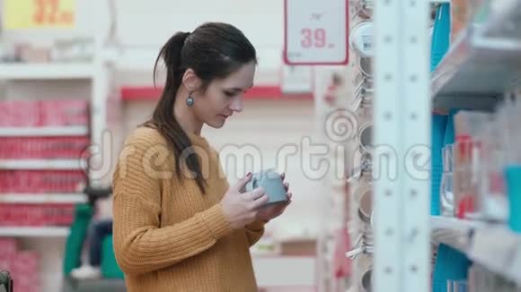 年轻漂亮的女人站在架子前拿着杯子拿着一个试着在超市里选择一些东西4K视频的预览图