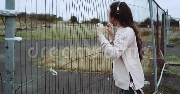 金属栅栏附近穿着粉红色背心的年轻女孩的肖像女孩在耳机里听音乐一边思考视频的预览图