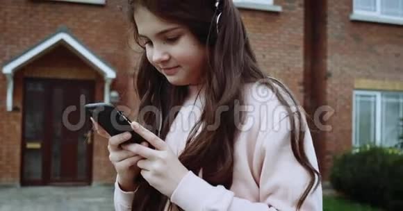 戴着耳机坐在家前院的漂亮女孩女孩用手机短信与乡间别墅视频的预览图