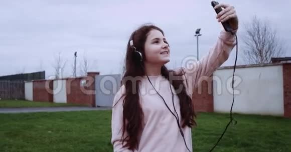 穿着粉色背心戴着耳机在砖围栏前自拍的漂亮女孩的肖像红色史诗相机在4K拍摄视频的预览图