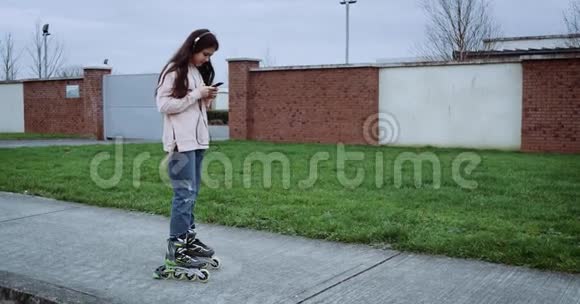 年轻可爱的女孩有着美丽的头发和耳机坐在绿草如茵的乡村街道上女孩在智能手机上发短信视频的预览图