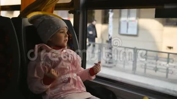 小金发女孩穿着粉红色的夹克孩子坐在公共汽车车厢里坐在她的座位上看着玻璃视频的预览图