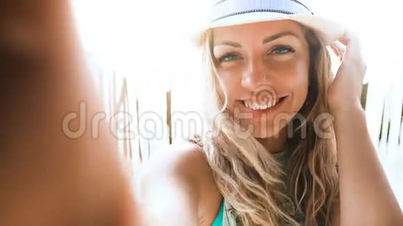 美丽的金发女郎戴着帽子在日落时分在沙滩上自拍视频的预览图