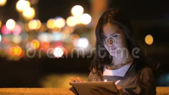 漂亮的年轻女人用平板电脑看着镜头微笑眨眼抚摸她的头发夜晚的城市模糊的灯光缓慢的视频的预览图