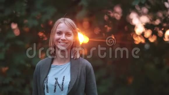 年轻美丽的女人站在公园的夕阳下微笑着笑着看着相机视频的预览图