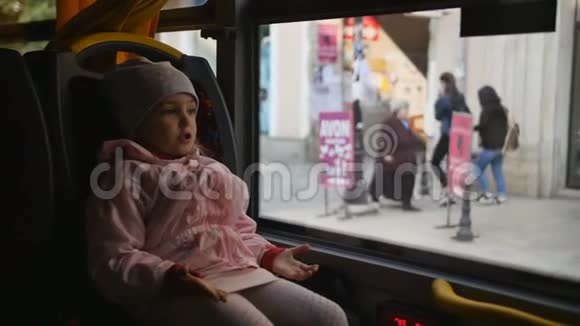 小金发女孩穿着粉红色的夹克孩子坐在公共汽车车厢里坐在她的座位上看着玻璃视频的预览图