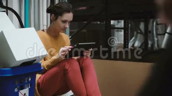 年轻女子坐在仓库里用平板电脑检查货物清单有手推车的人走到摄像机前面4K视频的预览图