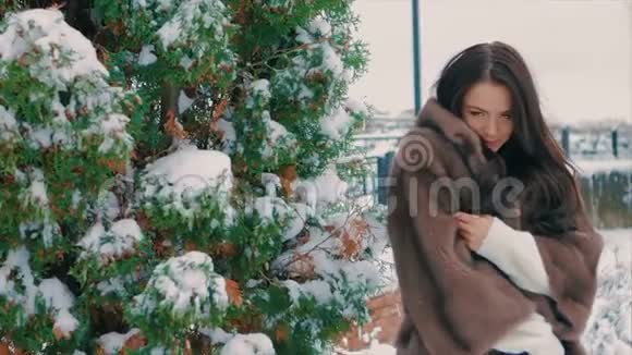 冬天在白雪覆盖的树木附近穿着棕色毛皮外套的时髦女孩慢动作视频的预览图
