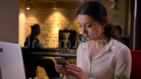 年轻的白种人女性办公室工作人员使用手机坐在工作场所笔记本电脑前的特写照片视频的预览图