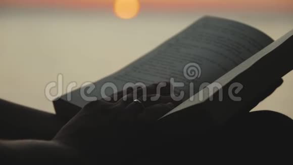 一个年轻女人坐在河边在日落时读一本书的剪影特写慢动作视频的预览图