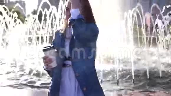 一个漂亮时髦的女孩站在喷泉边阳光明媚从纸杯里喝咖啡视频的预览图