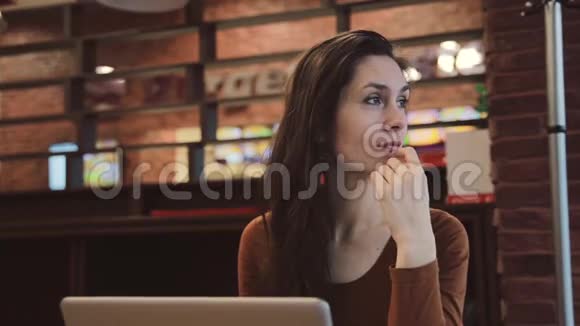 年轻的黑发女孩在咖啡馆里用笔记本电脑工作沉思地看着窗外视频的预览图