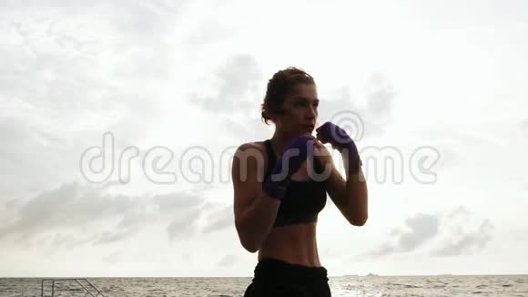 运动的年轻女子在海上背着太阳打拳击美丽的女拳击手在海滩上训练视频的预览图