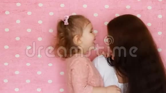 母女拥抱微笑玩得开心笑着亲吻年轻女子和她的孩子在粉色上的特写肖像视频的预览图