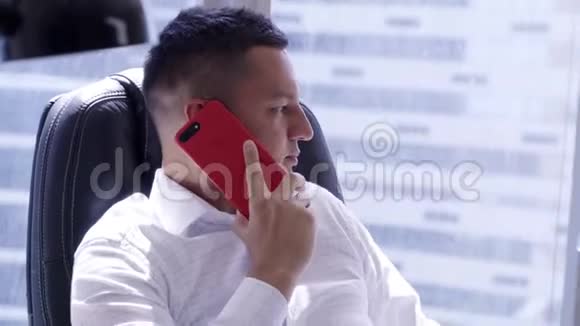 可爱成熟的黑发男子穿着白色衬衫在红色智能手机上讲话视频的预览图