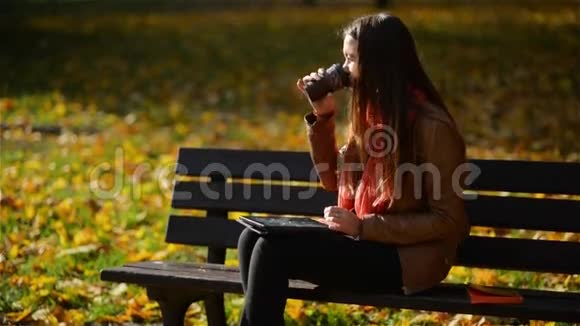 快乐的年轻学生女孩带着一张桌子和一个一次性咖啡杯坐在长凳上在秋天用笔记本电脑视频的预览图