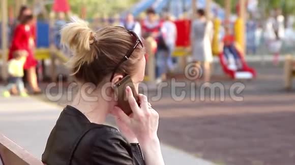 你的女人用手机坐在长凳公园里戴着时尚的酷墨镜妈妈等着她的女儿而她却在玩耍视频的预览图
