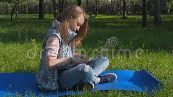 一个女孩坐在公园里拿着笔记本电脑一个十几岁的女孩坐在地毯上呼吸新鲜空气阳光明媚的一天一个带着笔记本电脑的女孩视频的预览图