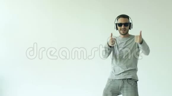 戴着太阳镜和耳机跳舞在白色背景下听音乐的快乐年轻人的慢动作视频的预览图