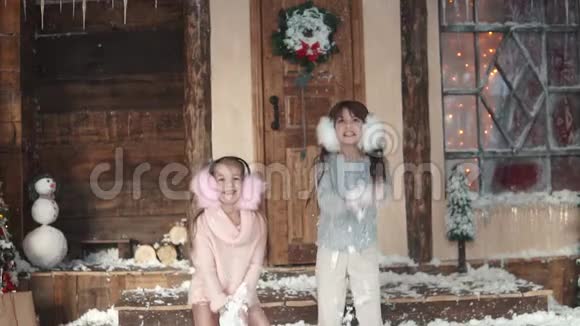 圣诞节或新年快乐的孩子们吐雪两个穿着毛皮耳机的小女孩两个小女孩在门廊上视频的预览图