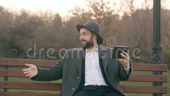 Hipser创意商人戴着帽子使用平板电脑在线视频聊天坐在公园的城市街凳上视频的预览图