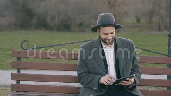 Hipser创意商业人士戴着帽子使用平板电脑休息时坐在公园的城市街凳上视频的预览图