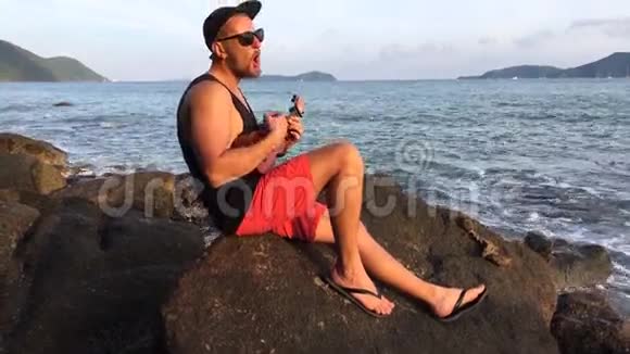 年轻英俊的胡须男在海滩岩石上唱歌和演奏四弦琴高清日落海景泰国普吉岛视频的预览图