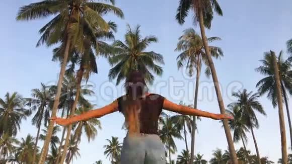 年轻快乐的女孩在椰子种植园里张开双手旅游妇女在棕榈树附近跳舞高清慢镜头视频的预览图