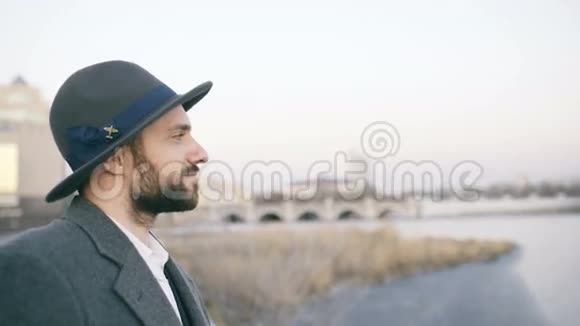 站在河边时戴着帽子和外套的留着胡子的年轻游客正在观看城市景观和白日梦视频的预览图