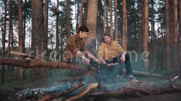 两个男人的朋友坐在树林里的原木上做烧烤聊天视频的预览图