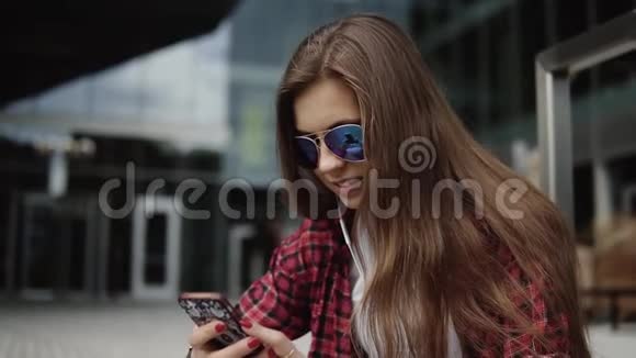 微笑的女人在她的智能手机上听音乐靠近玻璃般的现代办公室的台阶上视频的预览图