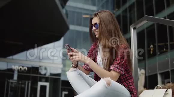 戴着时尚眼镜穿着休闲服装坐在台阶上听着iphone上音乐的满意女人微笑视频的预览图