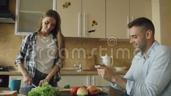 厨房里的漂亮情侣男人在智能手机上玩电子游戏而他的女朋友在做饭视频的预览图
