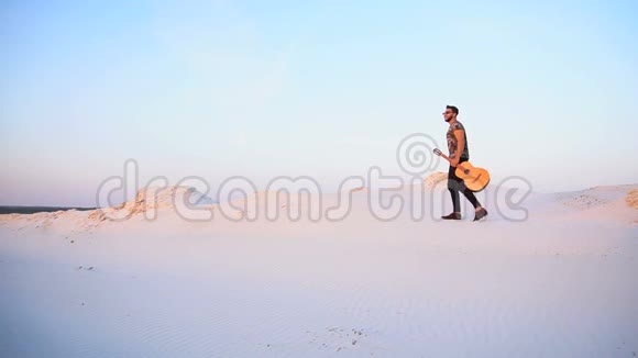 庄严的穆斯林年轻人手持吉他在夏季傍晚的露天日落时分穿过沙漠视频的预览图