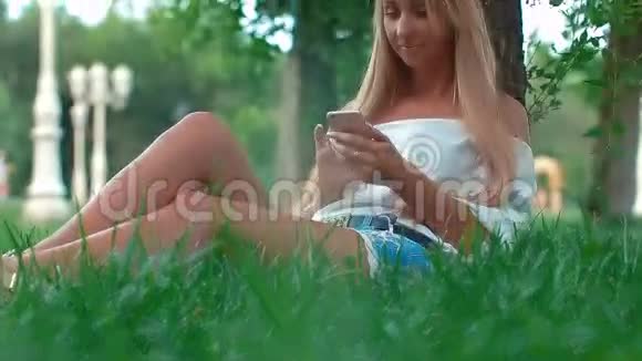 年轻女孩拿着智能手机坐在城市公园的绿草上女孩穿着一件白色的t恤视频的预览图