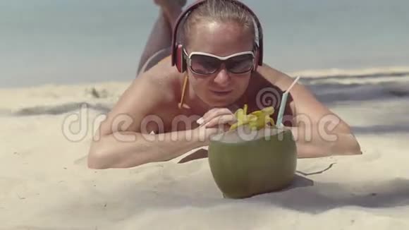 戴着太阳镜和耳机的女孩躺在海边的沙滩上看着椰子视频的预览图