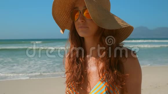穿着比基尼戴着墨镜和帽子站在海滩上的美丽的白种人年轻女子视频的预览图