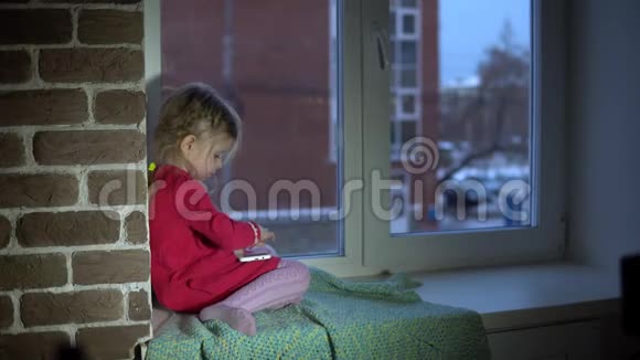 一个穿着粉红色裙子的小可爱女孩坐在窗台上用智能手机视频的预览图