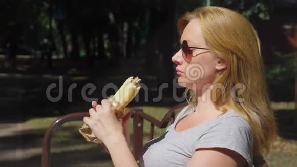 戴太阳镜的女孩愉快地吃热狗坐在城市公园的长凳上4k慢动作视频的预览图