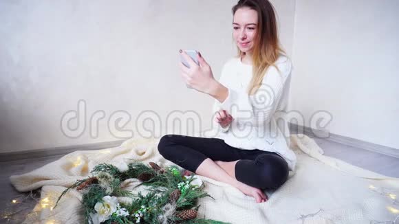 迷人的年轻女性用现代手机与朋友用视频电话交流坐在圣诞节旁边的地板上视频的预览图